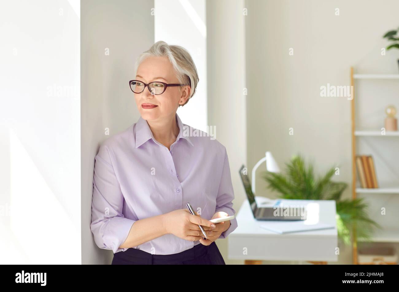Femme d'affaires souriante avec un téléphone portable entre les mains et regardant à distance par la fenêtre. Banque D'Images