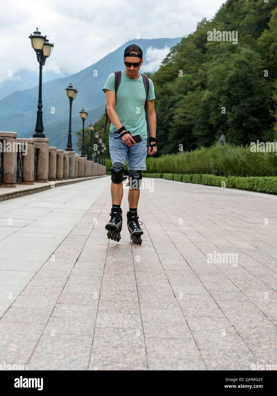 jeune homme en vêtements bleus en équipement de protection, sur des patins à roulettes le long d'un remblai sur fond de forêt et de montagnes en été Banque D'Images