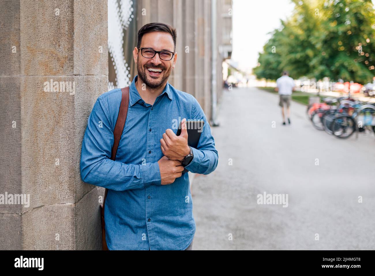 Portrait d'un entrepreneur joyeux tenant une tablette numérique. Le transport professionnel porte un sac pour ordinateur portable. Il est debout sur le trottoir de la ville. Banque D'Images