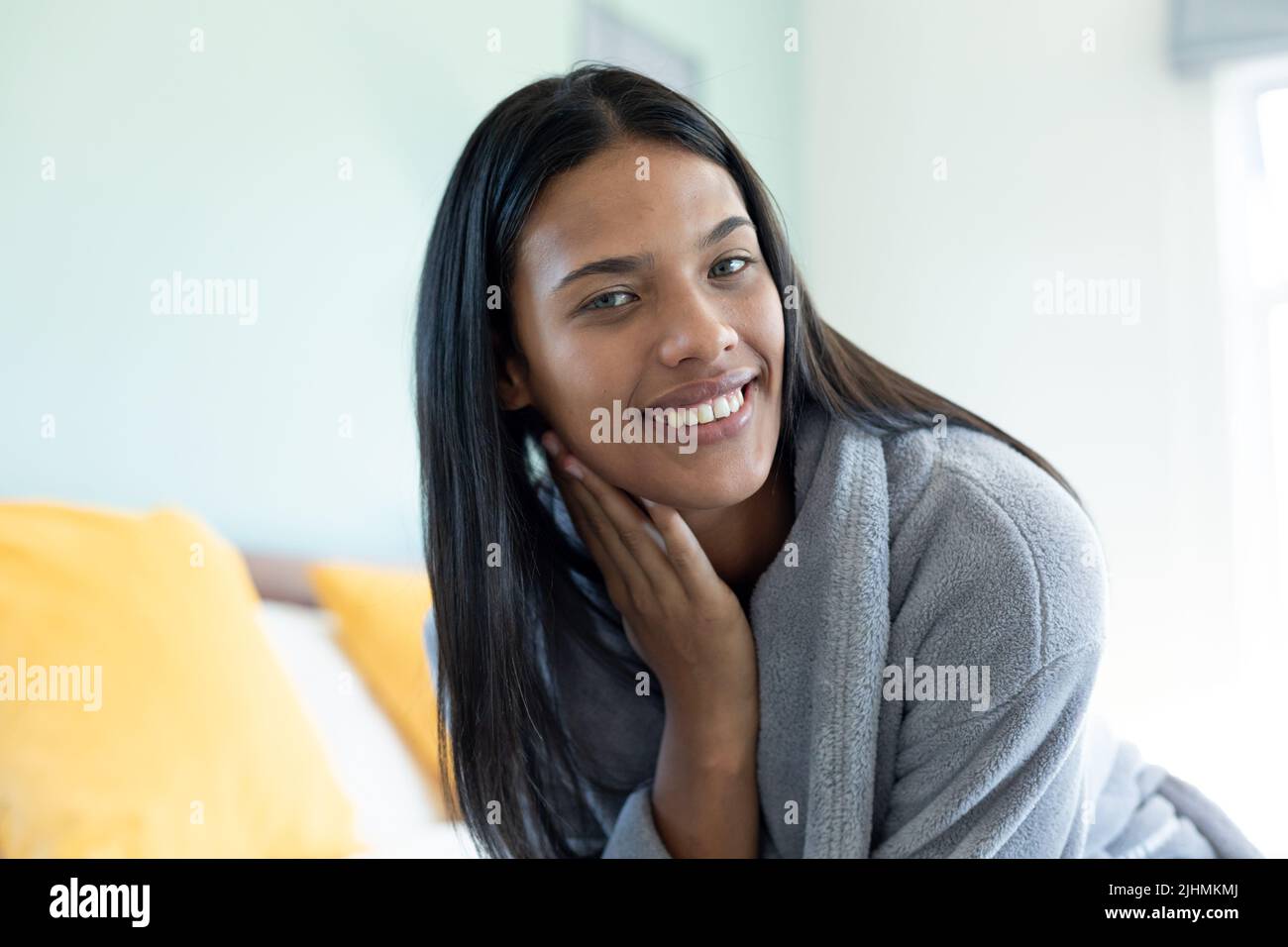 Portrait d'une jeune femme biraciale heureuse avec cheveux longs droits portant un peignoir contre le mur à la maison Banque D'Images