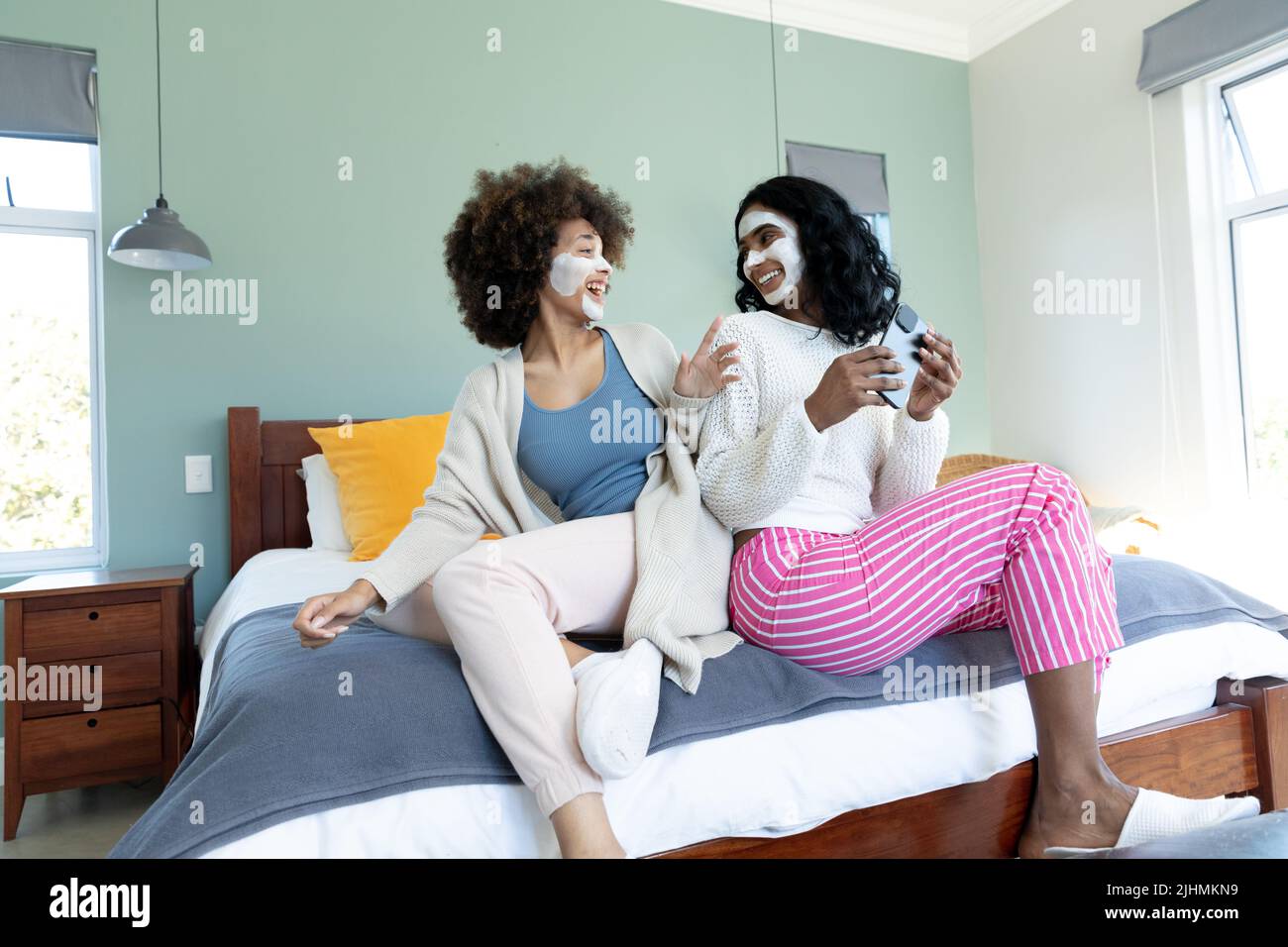 Jeunes amies biraciales gaies avec des crèmes faciales sur les visages regardant les uns les autres sur le lit Banque D'Images