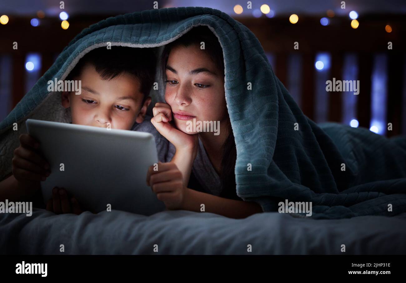 Bonne famille caucasienne avec deux enfants utilisant une tablette numérique couchée sous une couverture dans l'obscurité la nuit avec leur visage éclairé par l'écran de l'appareil Banque D'Images