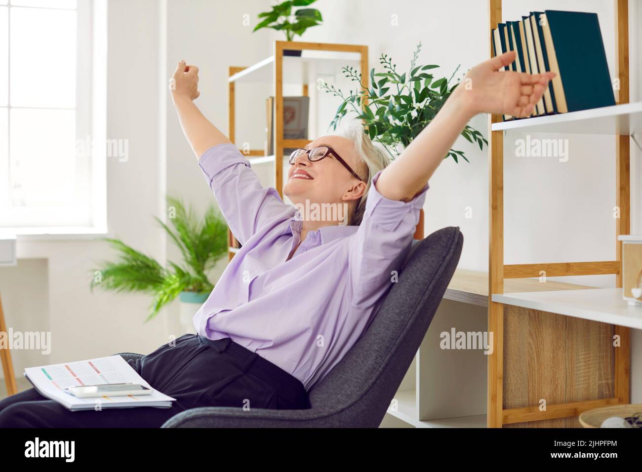 Heureuse dame sénior et détendue, assise sur une chaise et étirant ses bras après avoir terminé le travail Banque D'Images