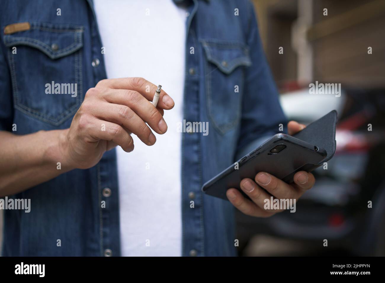 Gros plan. Dépendance malsaine fumer cigaret homme de lire les médias sociaux en utilisant son smartphone portant un Jean chemise et un t-shirt blanc debout à l'extérieur sur fond urbain ville. Aucune face visible. Banque D'Images