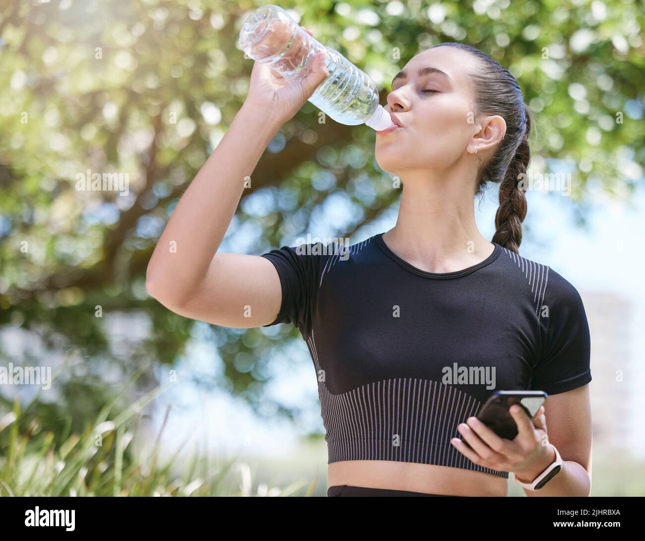Jeune athlète féminine prenant une pause et de l'eau potable à partir d'une bouteille et tenant un smartphone pendant une course et s'exerçant à l'extérieur pendant la journée Banque D'Images