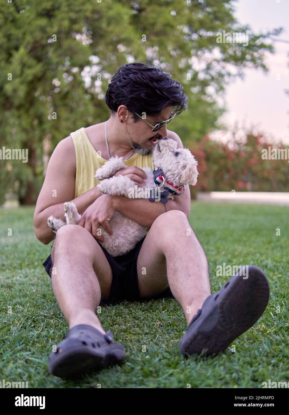 latino brun avec des lunettes de soleil assis sur la pelouse de son jardin jouant avec son animal de compagnie blanc jouet paodle un après-midi d'été. Mise au point sélective. Verti Banque D'Images