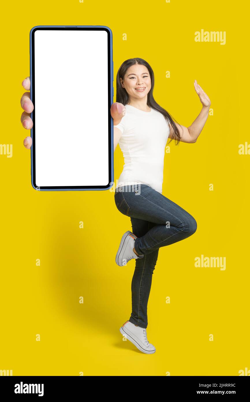 Bonne femme asiatique d'âge moyen montrant un smartphone avec écran blanc portant un t-shirt blanc et un Jean denim, publicité pour applications mobiles isolée sur fond jaune. Banque D'Images