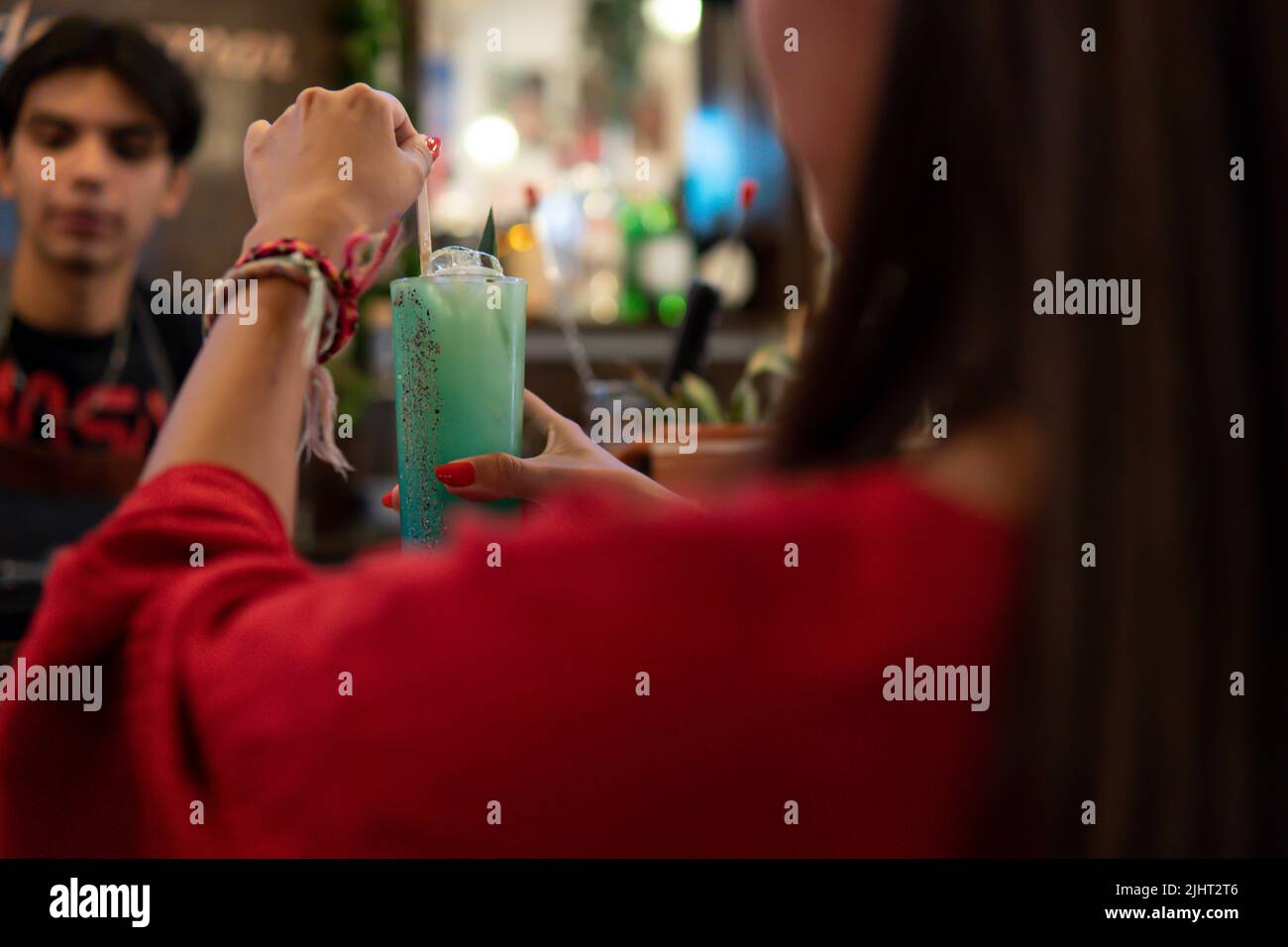 Fille aux cheveux bruns vêtue de rouge mélangeant la boisson que vient de servir le barman. Gros plan d'une fille avec une boisson alcoolisée dans un bar. Fille prenant un verre avec le serveur devant. Banque D'Images