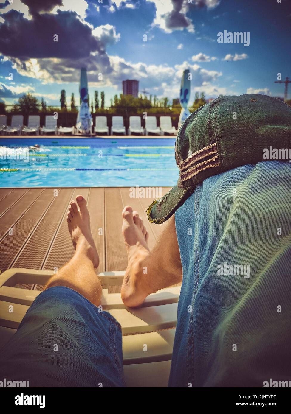 Les jambes des hommes dans un short en denim sur une chaise longue devant la piscine par temps ensoleillé. Casquette élégante sur le genou Banque D'Images Les jambes des hommes dans un short en denim sur une chaise longue devant la piscine par temps ensoleillé. Casquette élégante sur le genou Banque D'Images