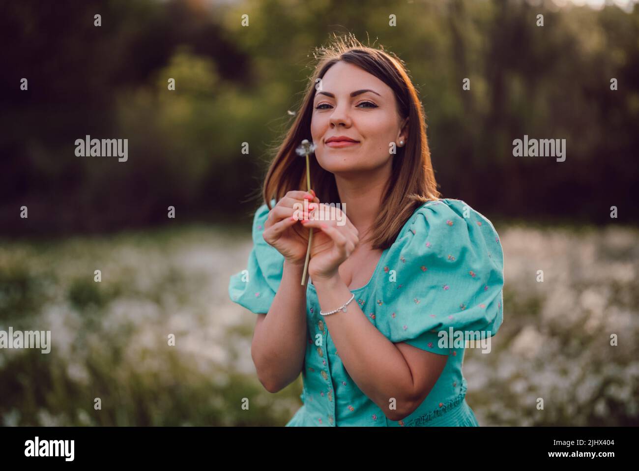 Femme souriante magnifique soufflant sur le pissenlit mûri dans le parc. Fille en robe turquoise rétro appréciant l'été à la campagne. Souhaitant, joie concept Banque D'Images