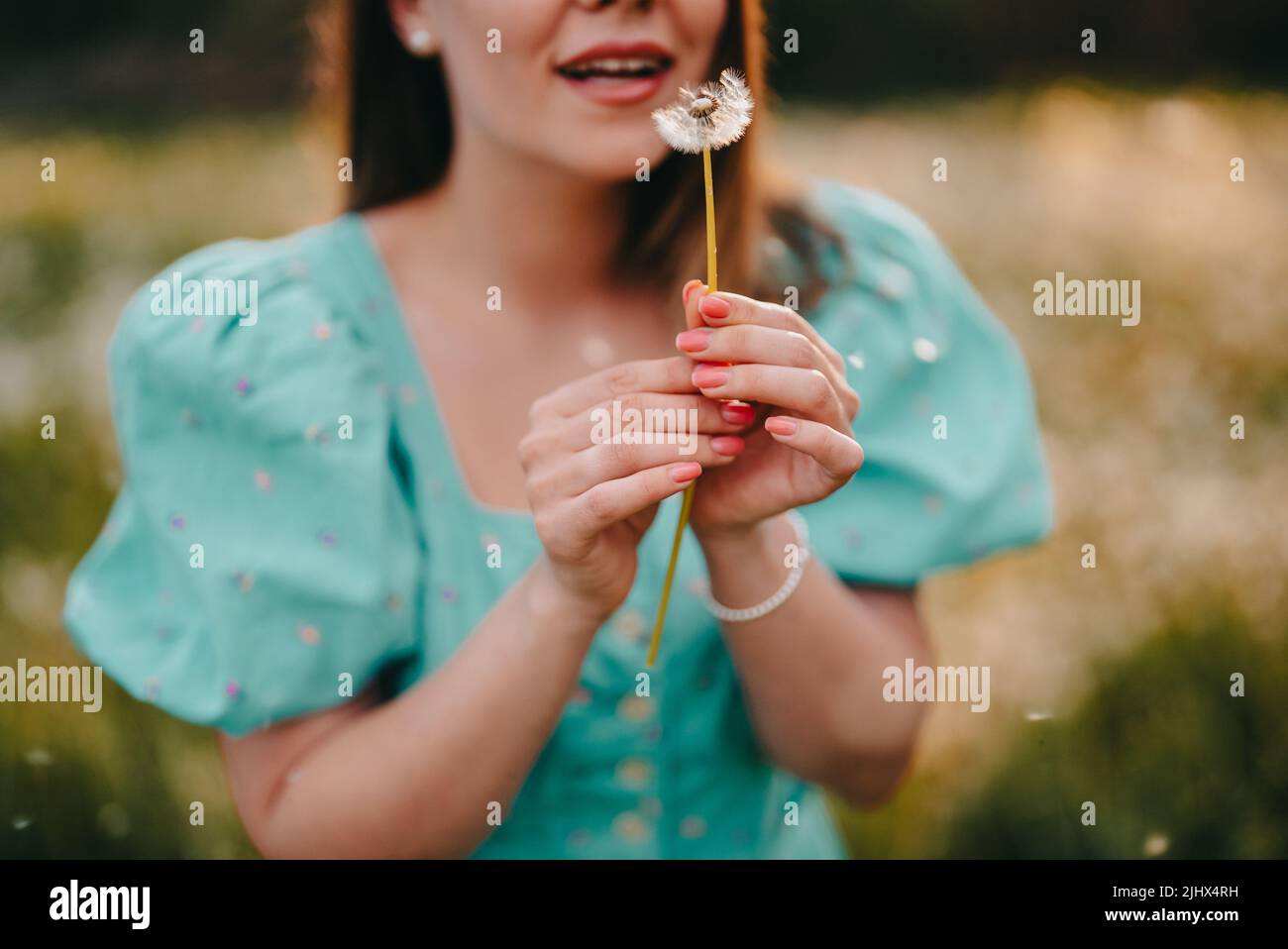 Femme souriante magnifique soufflant sur le pissenlit mûri dans le parc. Fille en robe turquoise rétro appréciant l'été à la campagne. Souhaitant, joie concept Banque D'Images