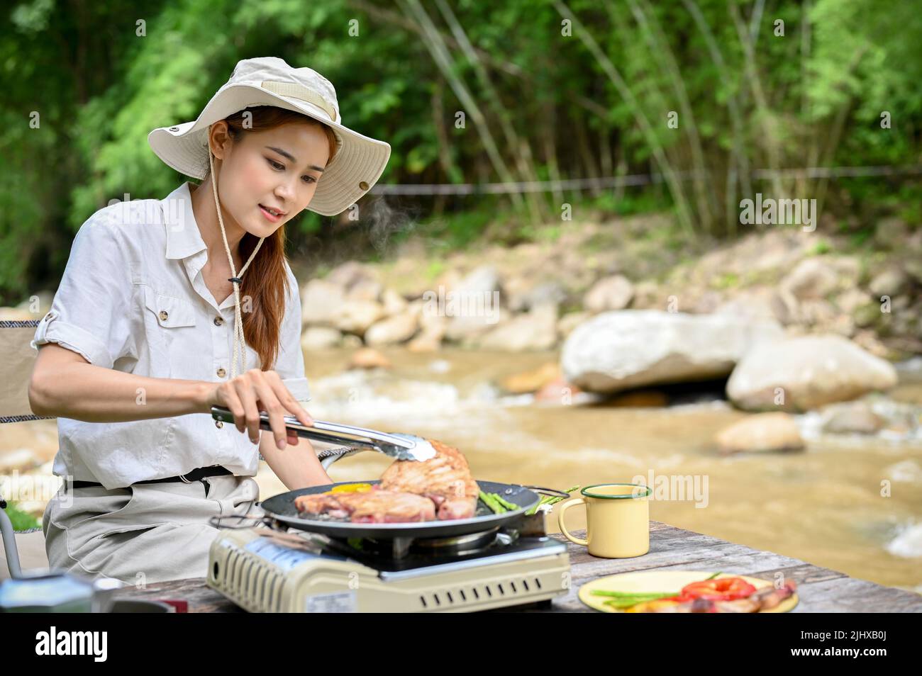 Charmante femme asiatique de camping-car millénaire grill quelques steaks de porc-hache sur un poêle de pique-nique portatif extérieur, cuisant son petit déjeuner près de la rivière. Banque D'Images