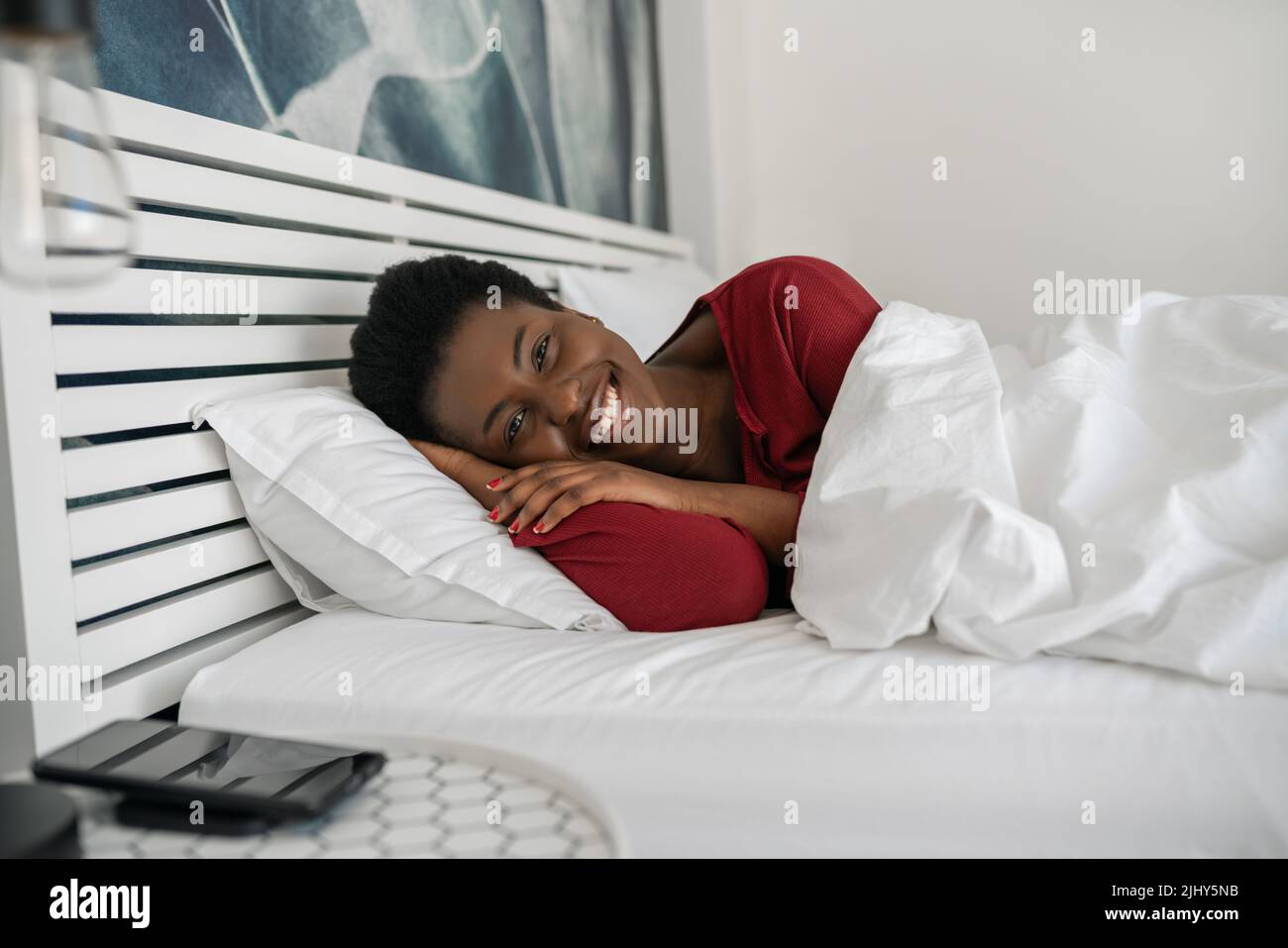 Belle femme africaine posée dans le lit, regardant dans l'appareil photo et souriant, posée confortablement sous la couette et sur les oreillers le matin, juste réveillé Banque D'Images