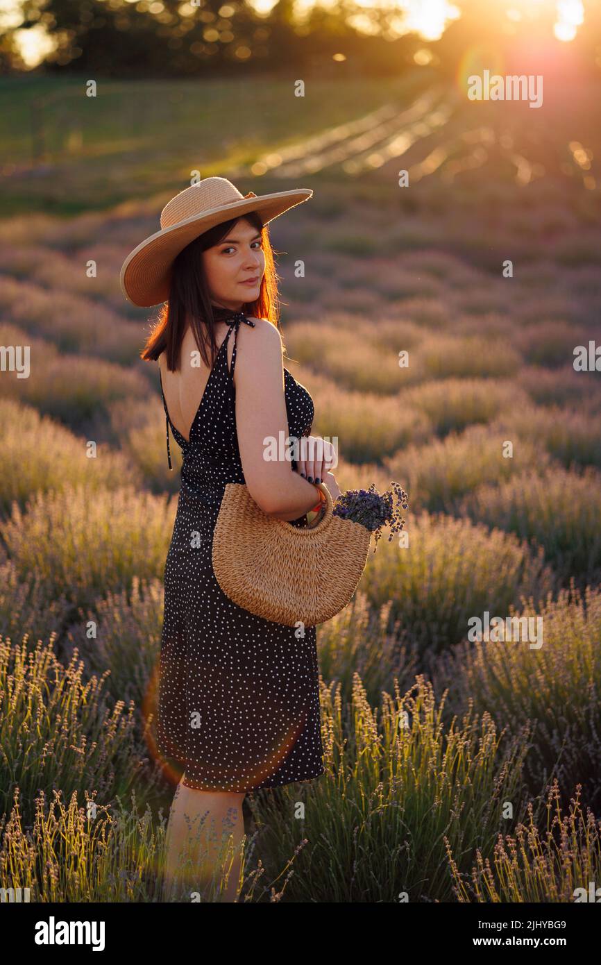 Jeune femme en robe et chapeau de paille debout dans un champ de lavande avec sac en osier Banque D'Images