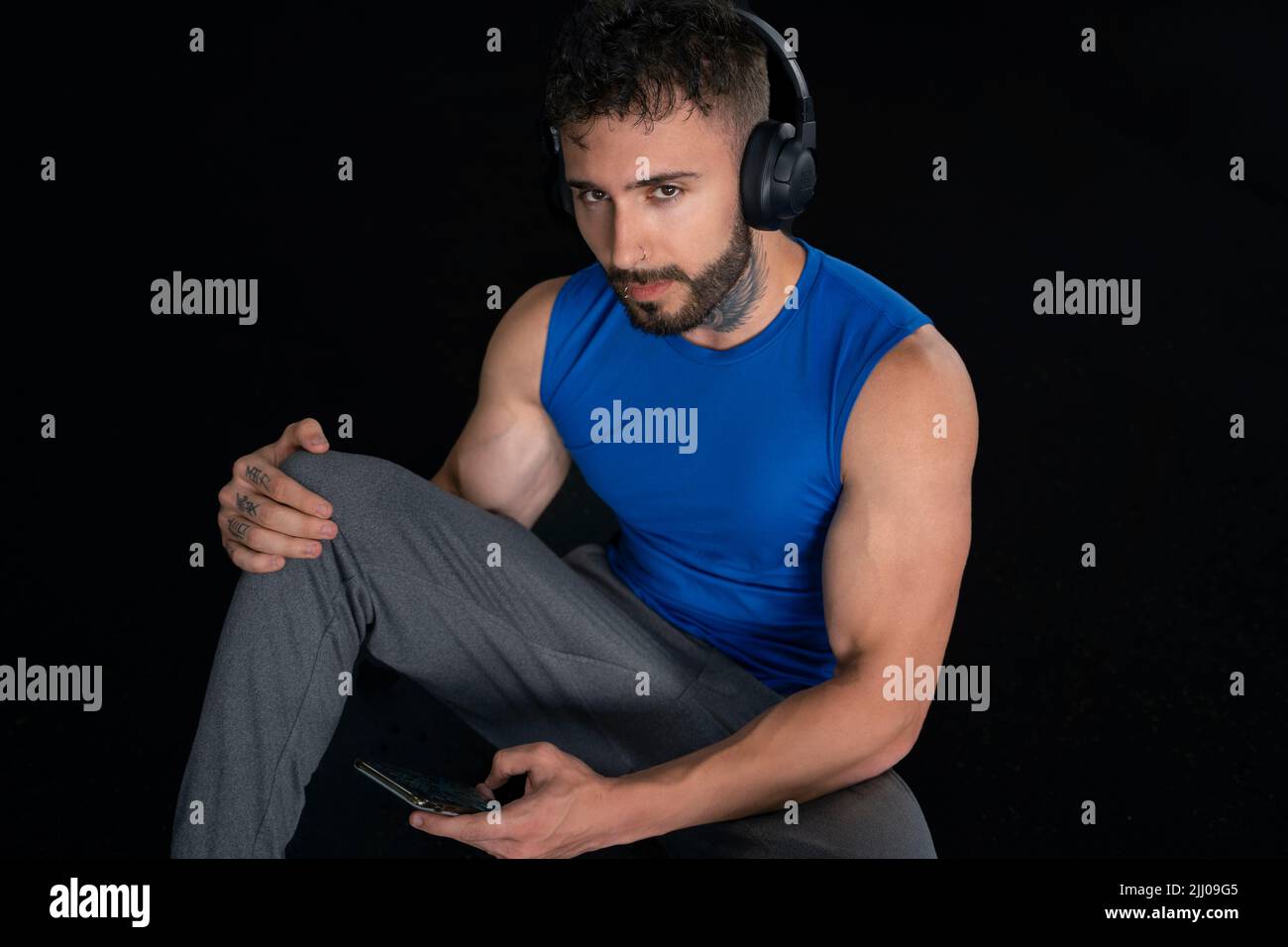 Beau homme avec tatouages dans son 20s dans la salle de gym avec téléphone et écouteurs, isolé sur fond noir Banque D'Images