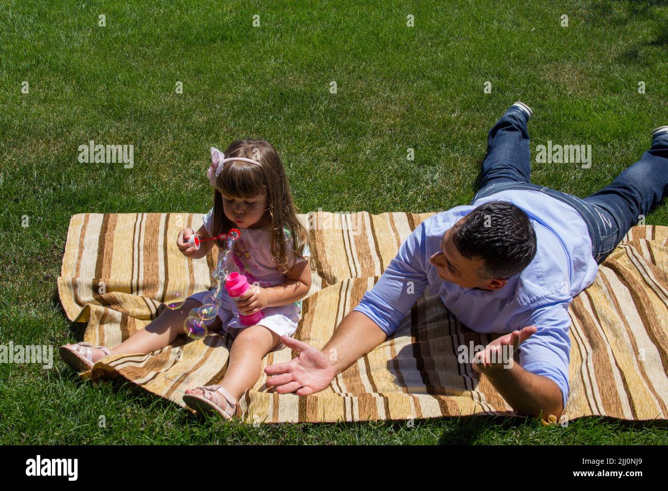 Image d'un jeune père assis dans un pré vert jouant avec sa fille faisant des bulles de savon. Référence au lien entre le père et la fille Banque D'Images