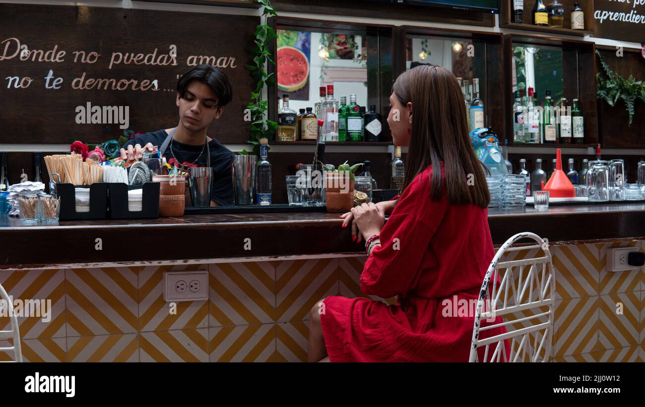 Fille hispanique en robe rouge assise au comptoir du bar pour commander une boisson auprès du barman. Serveur servant une fille latine. Bar avec l'esthétique traditionnelle avec beaucoup de plantes et de bouteilles d'alcool. Banque D'Images