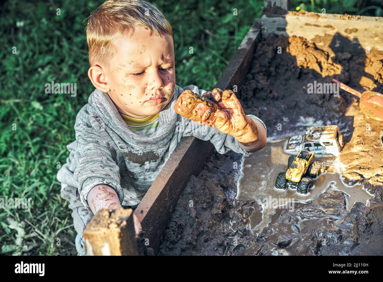 Mignon petit garçon sérieux avec le visage sale et les vêtements jouant avec des voitures jouets dans le bac à sable près de maison rurale en bois en été dans la campagne Banque D'Images