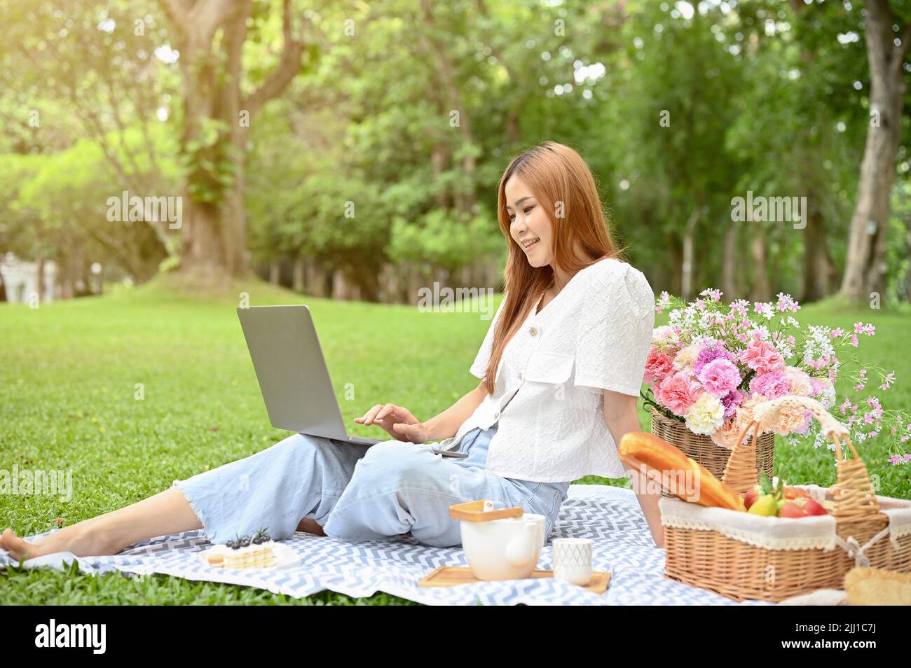 Charmante jeune femme asiatique ou étudiante pique-nique seule dans le parc verdoyant, assise sur son linge de pique-nique et utilisant un ordinateur portable pour gérer Banque D'Images