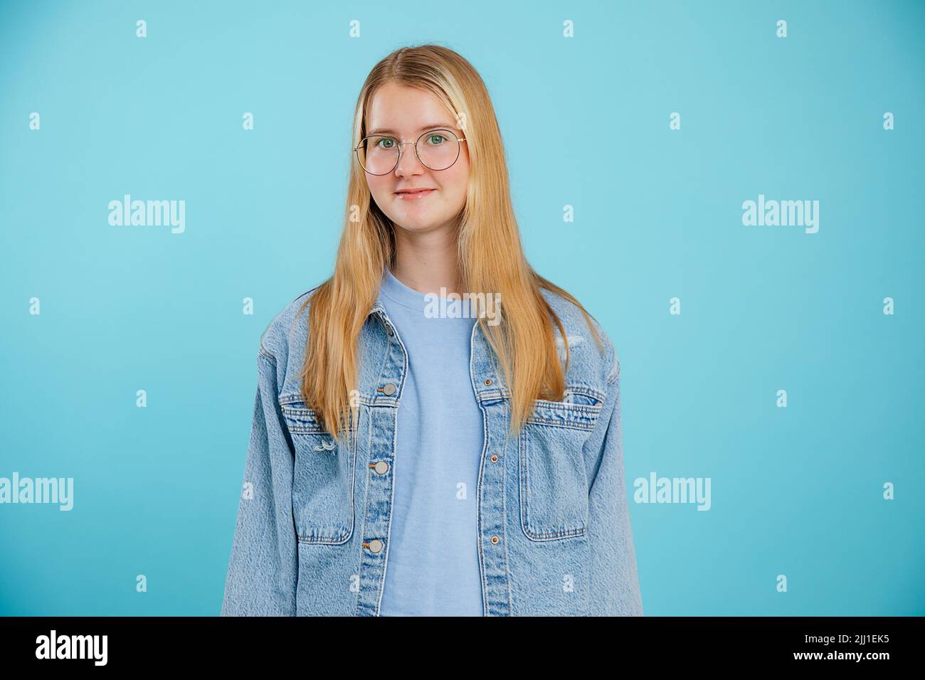 Jeune femme blonde calme positive, étudiante regardant un appareil photo dans des lunettes et veste de Jean dans un fond de studio bleu Banque D'Images