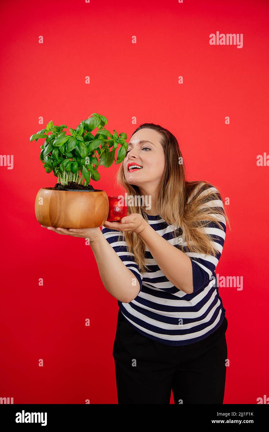 Vertical souriant long-cheveux plump femme dans une chemise à rayures tenant et odeur de basilic pot de plante et de pomme fraîche. Soins de santé Banque D'Images