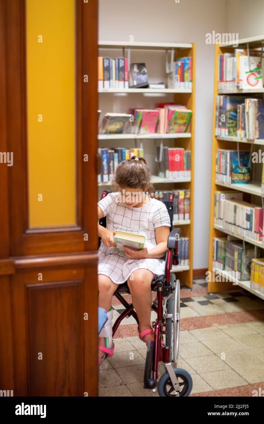 Une petite fille est assise dans un fauteuil roulant et lit un livre dans la bibliothèque. La vie et l'éducation des enfants ayant des besoins spéciaux. Enfant handicapé heureux Banque D'Images