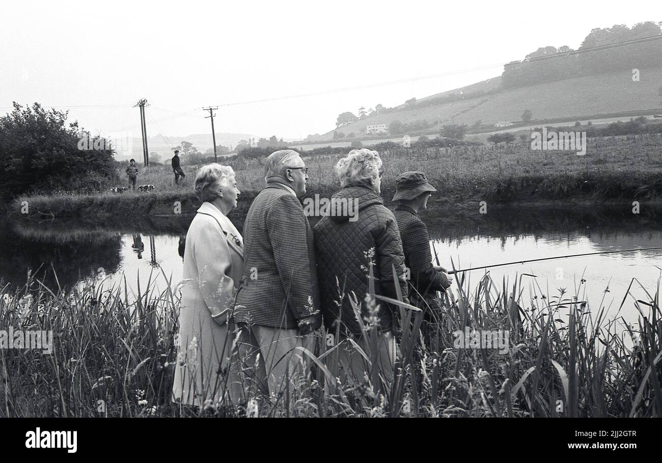 1960s, historique, trois personnes se tenant sur une rive derrière un pêcheur qui le regarde pêcher dans la rivière, Tewkesbury, Angleterre, Royaume-Uni. Banque D'Images