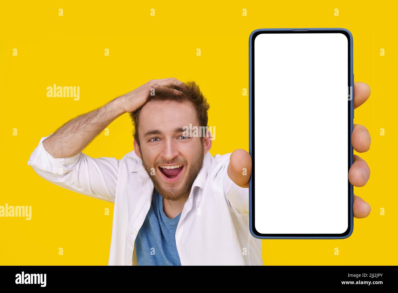 Beau jeune homme excité souriant émotionnellement touchant sa tête montrant un immense smartphone avec écran blanc. Mise en place de maquettes publicitaires d'applications mobiles. Jeune homme avec téléphone. Banque D'Images