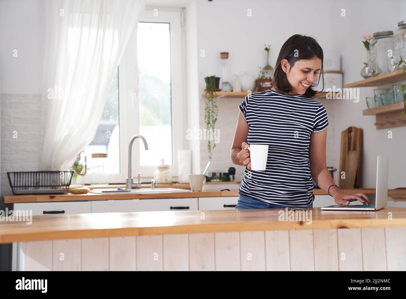 Une femme dans la cuisine à l'ordinateur avec un sourire. Une femme tape sur un ordinateur, travaille à domicile ou communique sur les réseaux sociaux. Une femme boit du thé et tient une tasse dans sa main. Photo de haute qualité Banque D'Images
