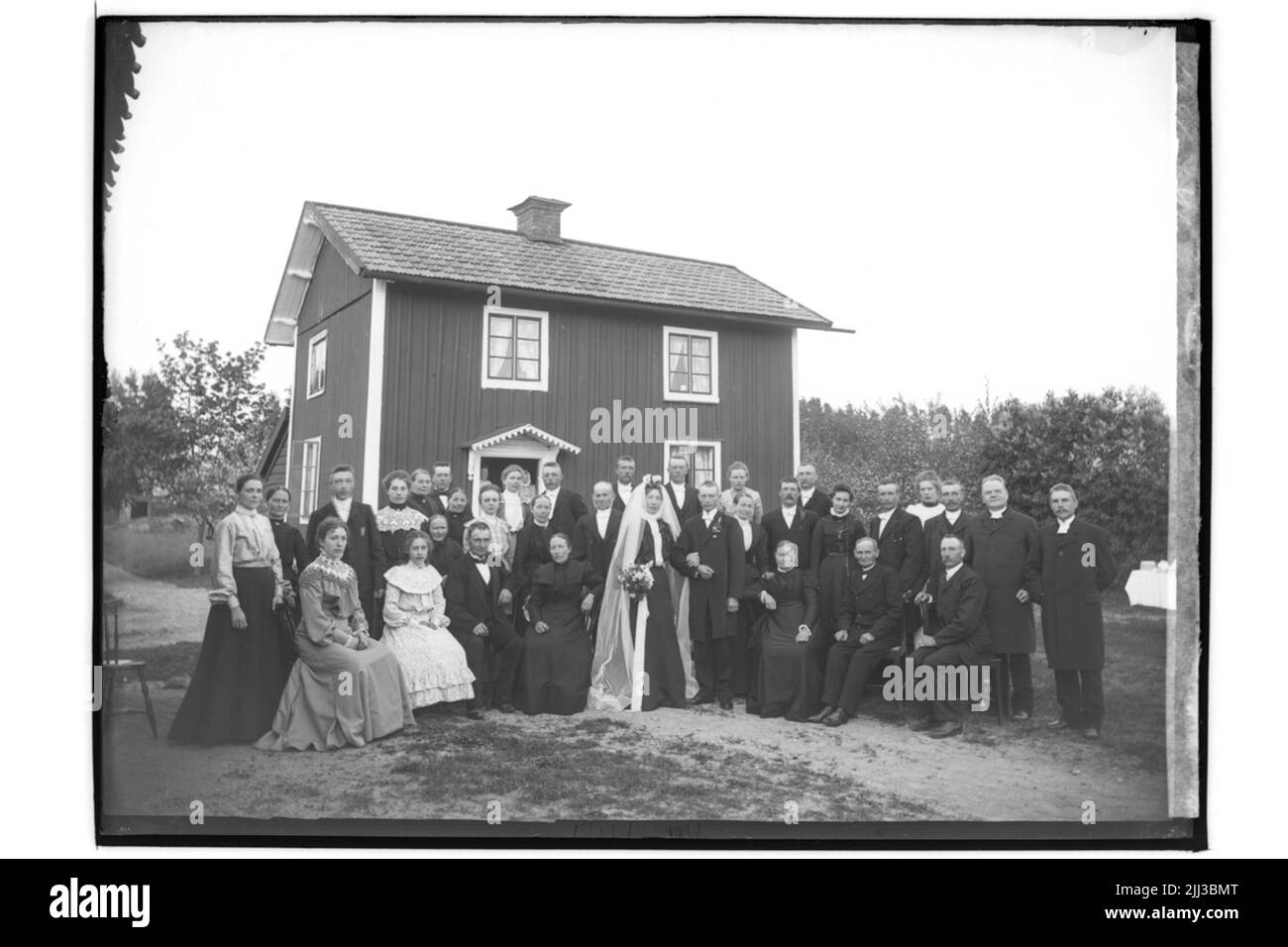 Mariage, 34 personnes. La mariée en robe noire, voile blanc, couronne. Le marié dans la cabane. Banque D'Images