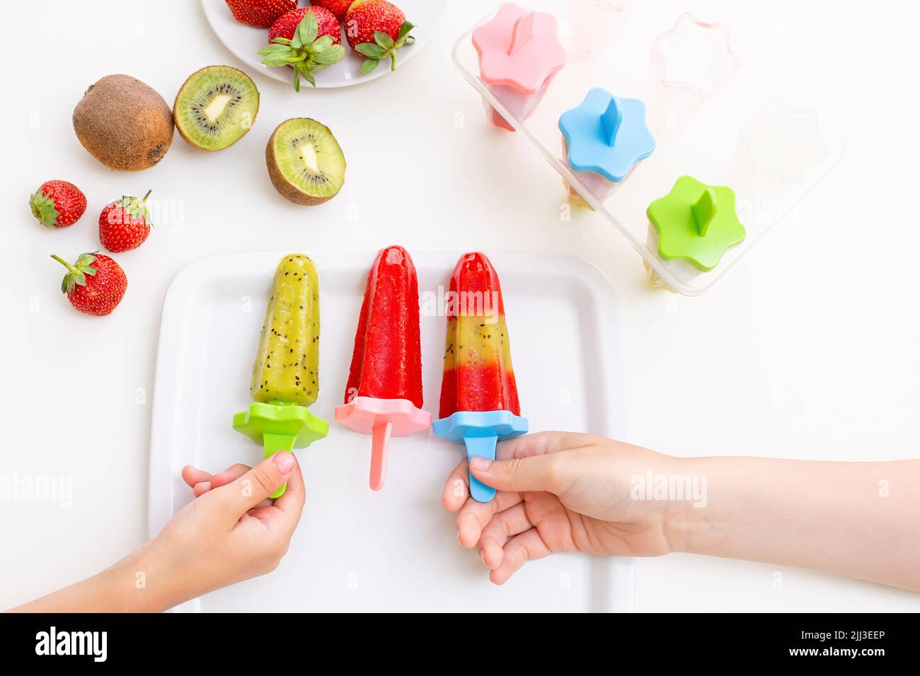 Deux mains pour enfants, prenez de la glace aux kiwis et aux fraises multicolores faits maison dans une assiette carrée blanche Banque D'Images