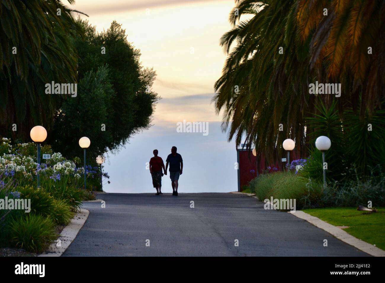 Deux amoureux prennent une promenade au coucher du soleil sur une route ou une allée tranquille avec des feux de rue et des palmiers les encadrent à Rutherglen Banque D'Images