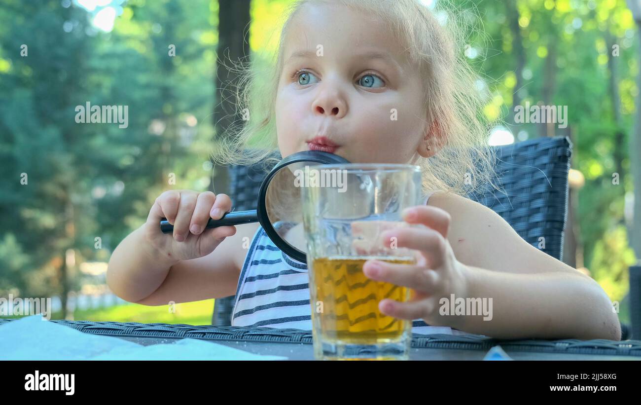 La petite fille regarde dans l'objectif autour. Gros plan d'une fille blonde étudiant le monde autour d'elle la regardant à travers la loupe tout en étant assise Banque D'Images