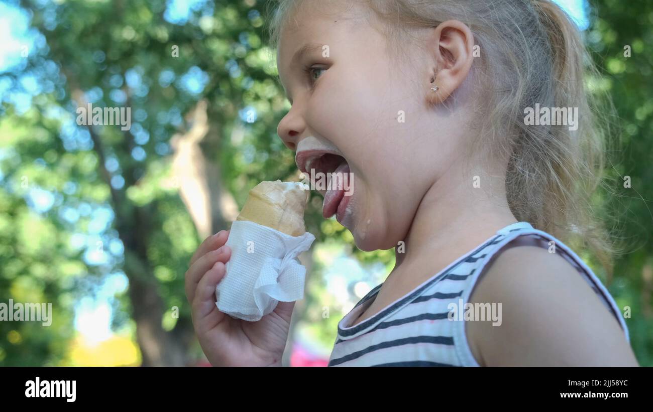 Une petite fille mignonne mange de la glace à l'extérieur. Portrait en gros plan d'une fille blonde assise sur le banc du parc et mangeant de la glace. Banque D'Images