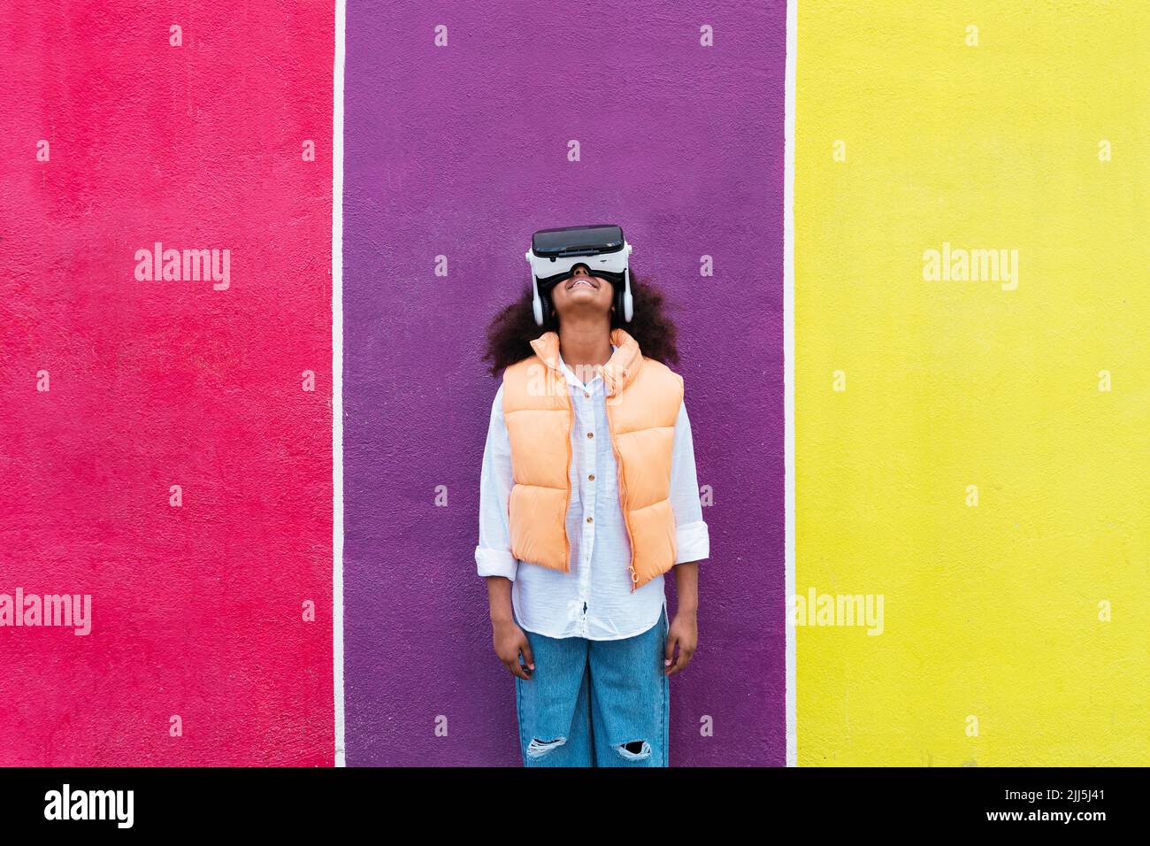 Fille souriante portant un simulateur de réalité virtuelle devant un mur multicolore Banque D'Images