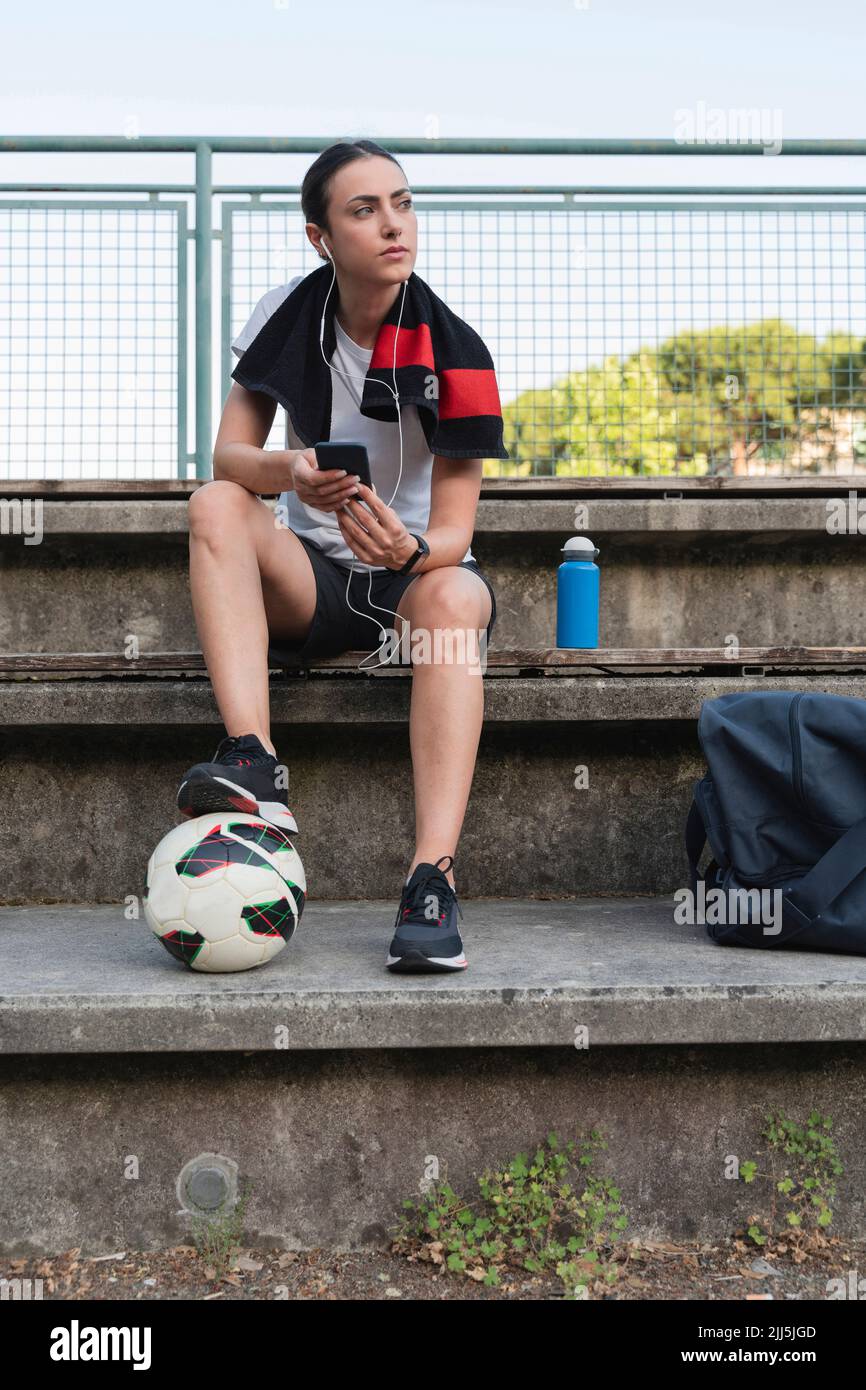 Jeune femme avec ballon de football à écouter de la musique à travers les écouteurs intra-auriculaires sur banc Banque D'Images