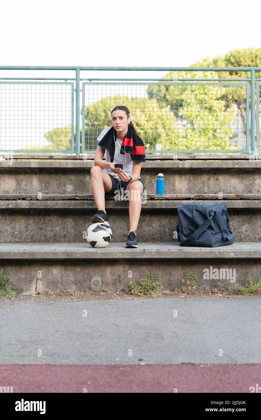 Femme écoutant de la musique avec un casque intra-auriculaire assis avec un ballon de football sur un banc Banque D'Images