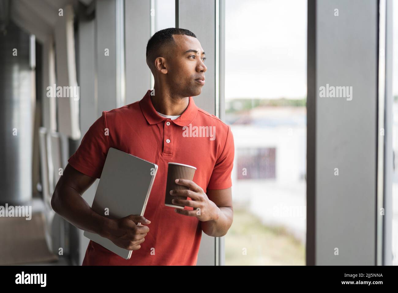 Homme d'affaires avec une tasse jetable et un ordinateur portable donnant par la fenêtre au bureau Banque D'Images
