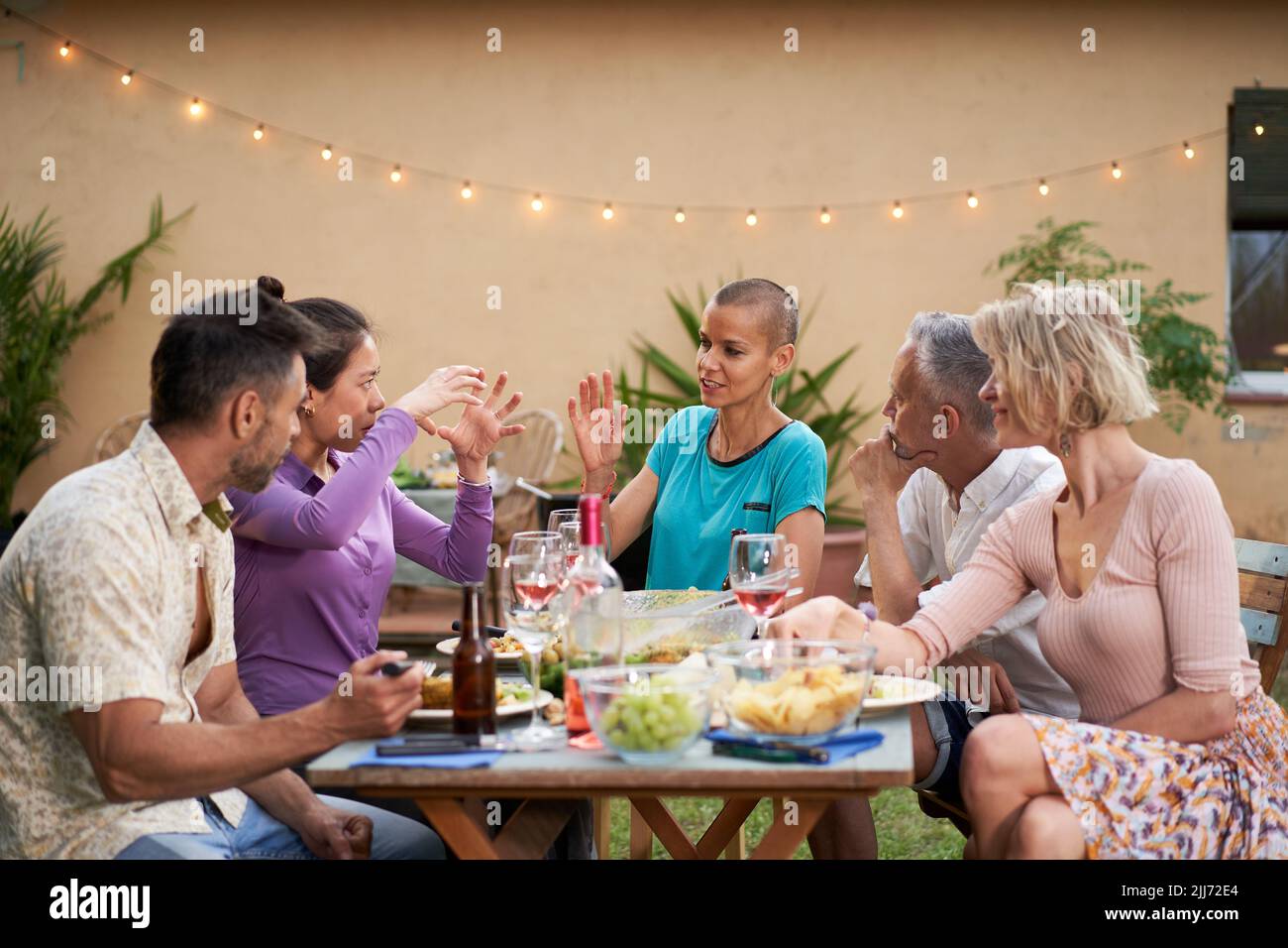 Groupe d'amis discutant pendant le déjeuner dans l'arrière-cour. Personnes d'âge moyen ayant une discussion à l'extérieur à la table tout en mangeant et en buvant Banque D'Images