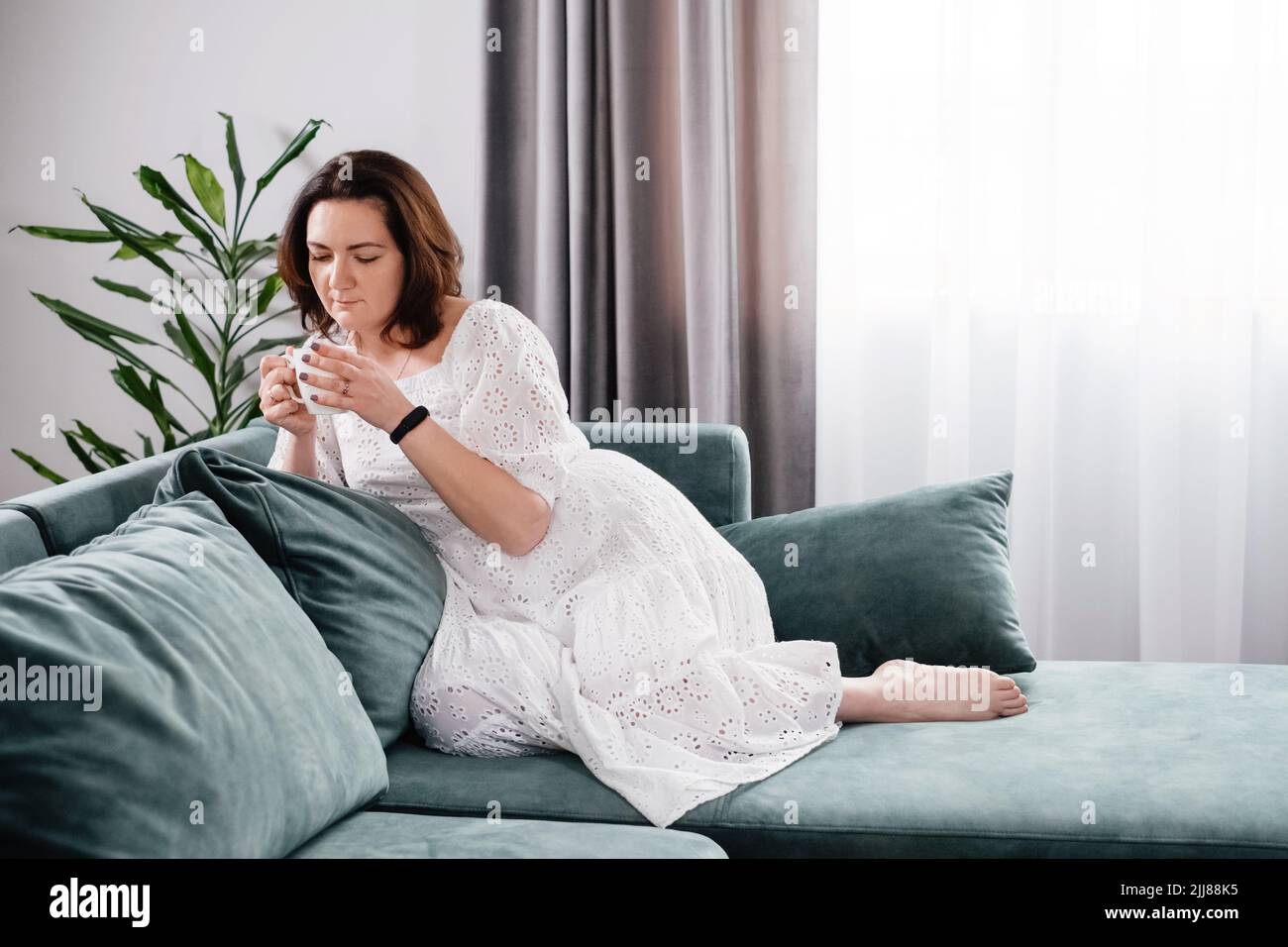 Une femme boit une tasse de café dans une atmosphère chaleureuse le matin. Vraie femme d'âge moyen plus grande en robe blanche se relaxant sur le canapé, moderne Banque D'Images