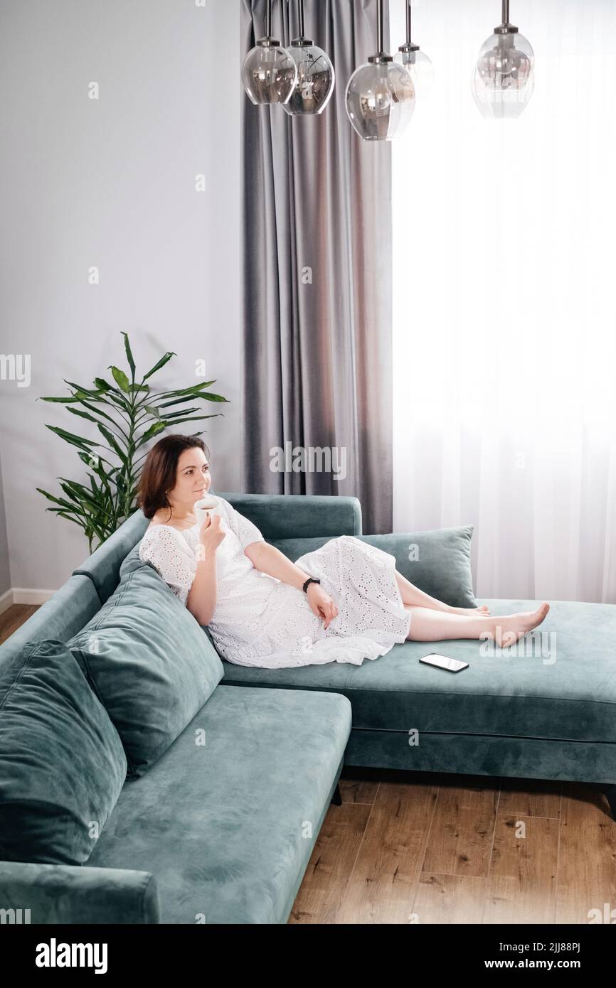 Une femme boit une tasse de café dans une atmosphère chaleureuse le matin. Vraie femme d'âge moyen plus grande en robe blanche se relaxant sur le canapé, moderne Banque D'Images