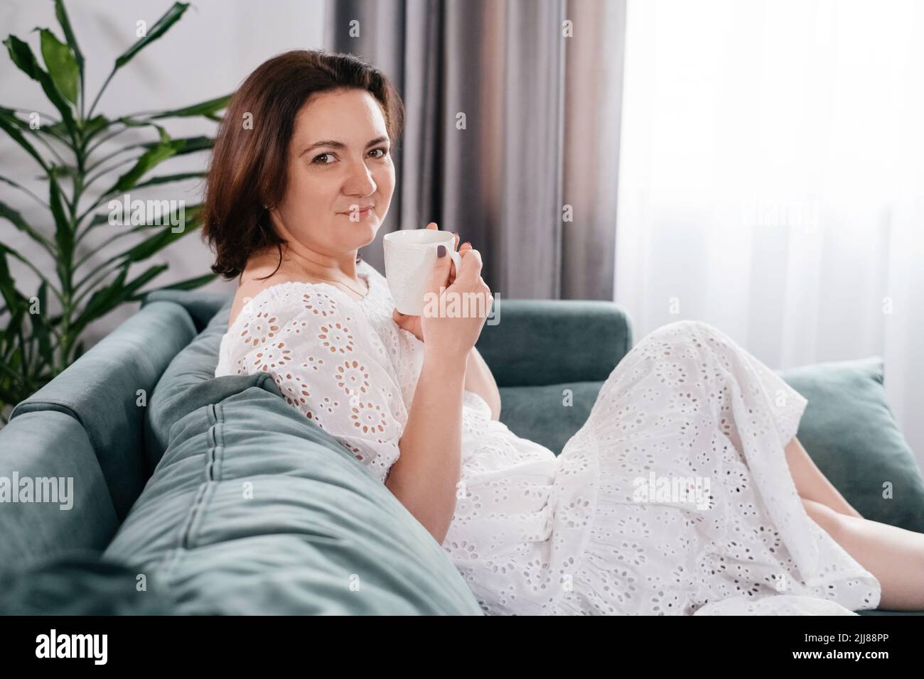 Une femme boit une tasse de café dans une atmosphère chaleureuse le matin. Vraie femme d'âge moyen plus grande en robe blanche se relaxant sur le canapé, moderne Banque D'Images