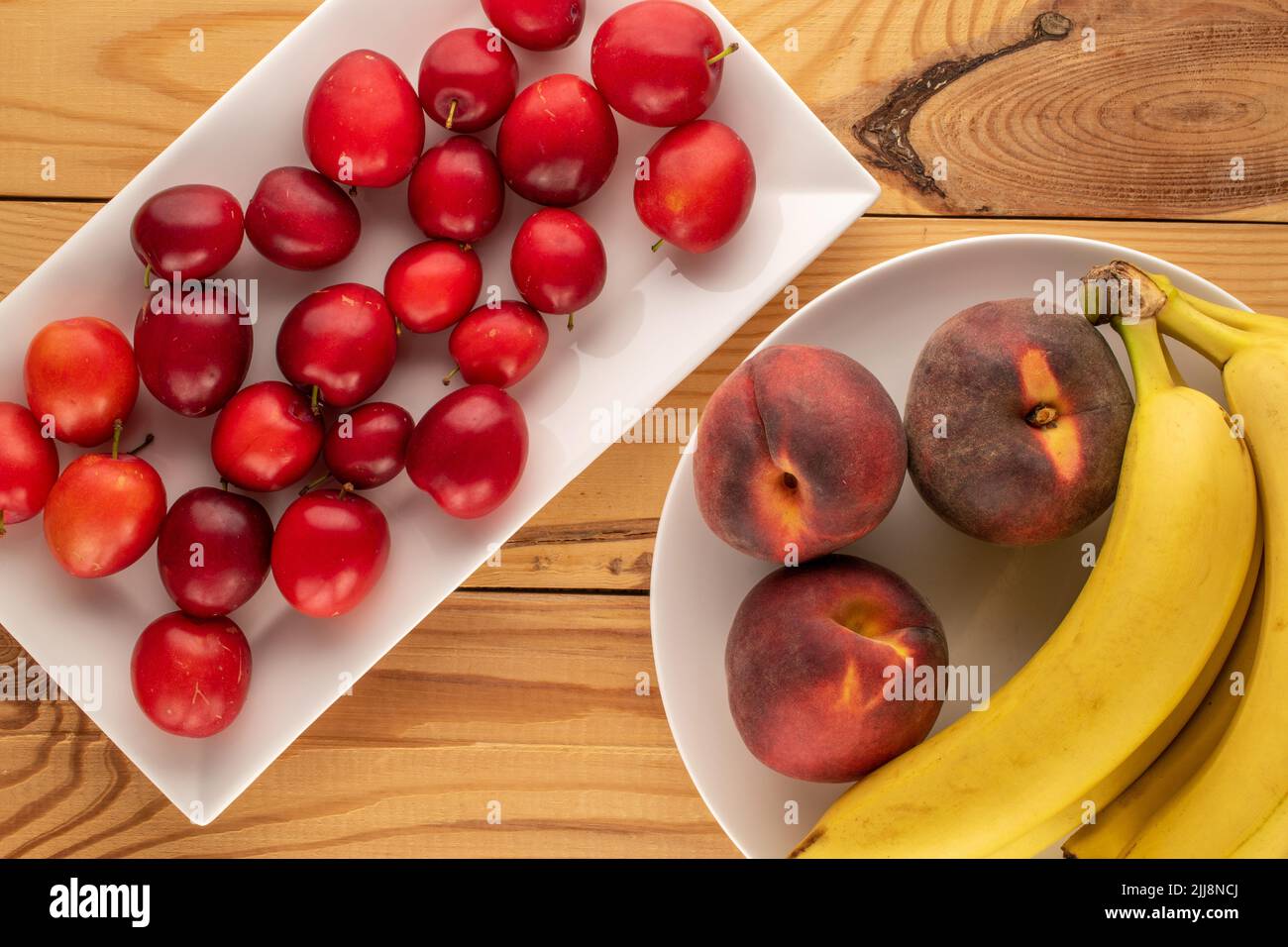 Plusieurs prunes de cerise sucrée, trois pêches et un bouquet de bananes avec des ustensiles en céramique blanche sur une table en bois, vue rapprochée, vue de dessus. Banque D'Images