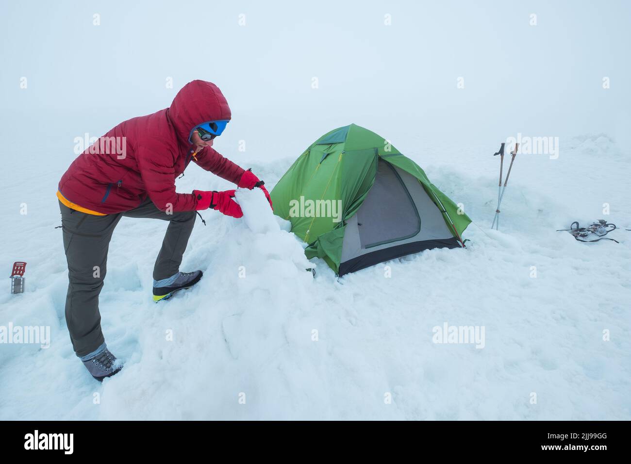 Homme souriant habillé rouge chaud alpinisme veste en bas faisant le mur de protection des briques de neige autour du camp tente sur le plateau de glacier 4200m sous Ka Banque D'Images