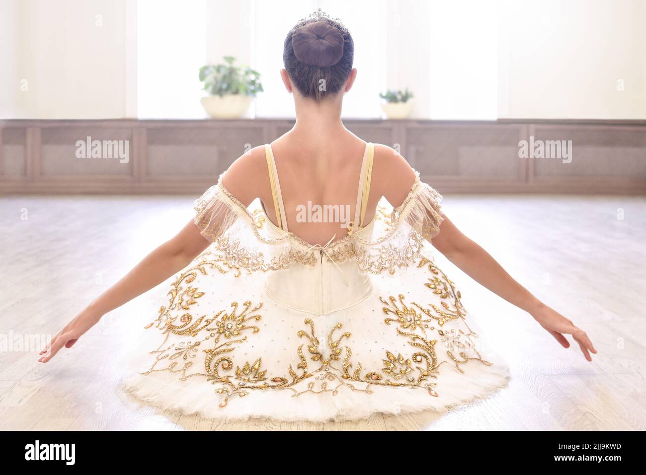 Vue arrière de la magnifique jeune danseuse de ballet en robe crème assise et se réchauffe les mains sur un plancher de bois dans le studio de ballet Banque D'Images
