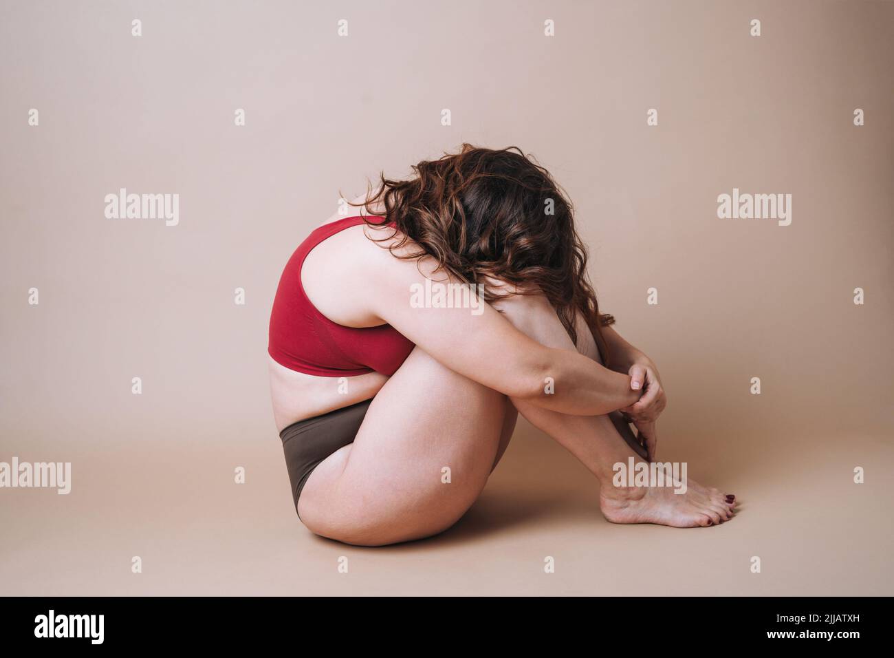 Jeune femme malheureuse plus la taille dans les sous-vêtements assis en position fermée sur fond beige Banque D'Images