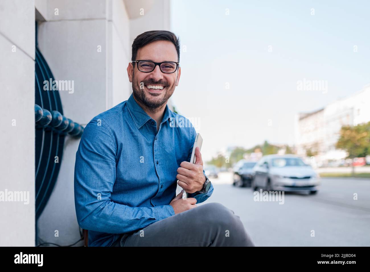 Portrait d'un jeune homme d'affaires souriant tenant un ordinateur portable. Un homme joyeux et professionnel se rendant dans des coffals est assis contre le bâtiment. Il se déplace dans t Banque D'Images