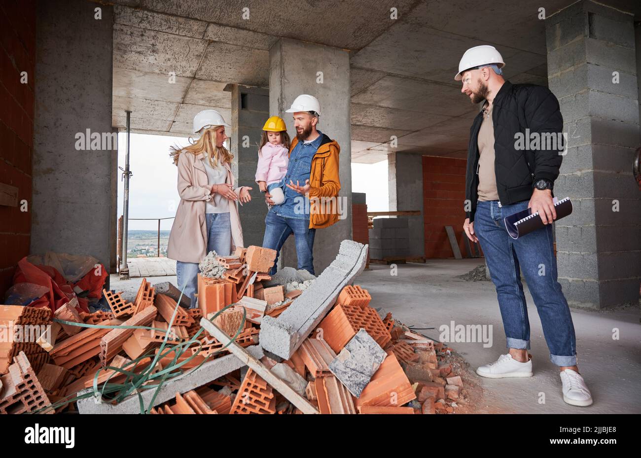 Homme et femme avec enfant debout à l'intérieur de l'immeuble d'appartements en construction et parlant avec le constructeur. Famille futurs propriétaires discutant du processus de construction avec le travailleur de la construction. Banque D'Images