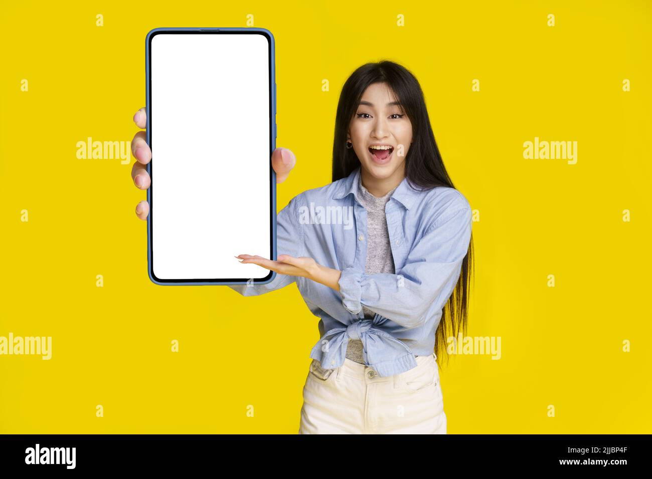 Grand smartphone avec écran blanc en main de la belle fille asiatique heureux de présenter une nouvelle application, jeu, gagner, isolé sur fond jaune. Placement de produits publicitaires pour applications mobiles. Banque D'Images