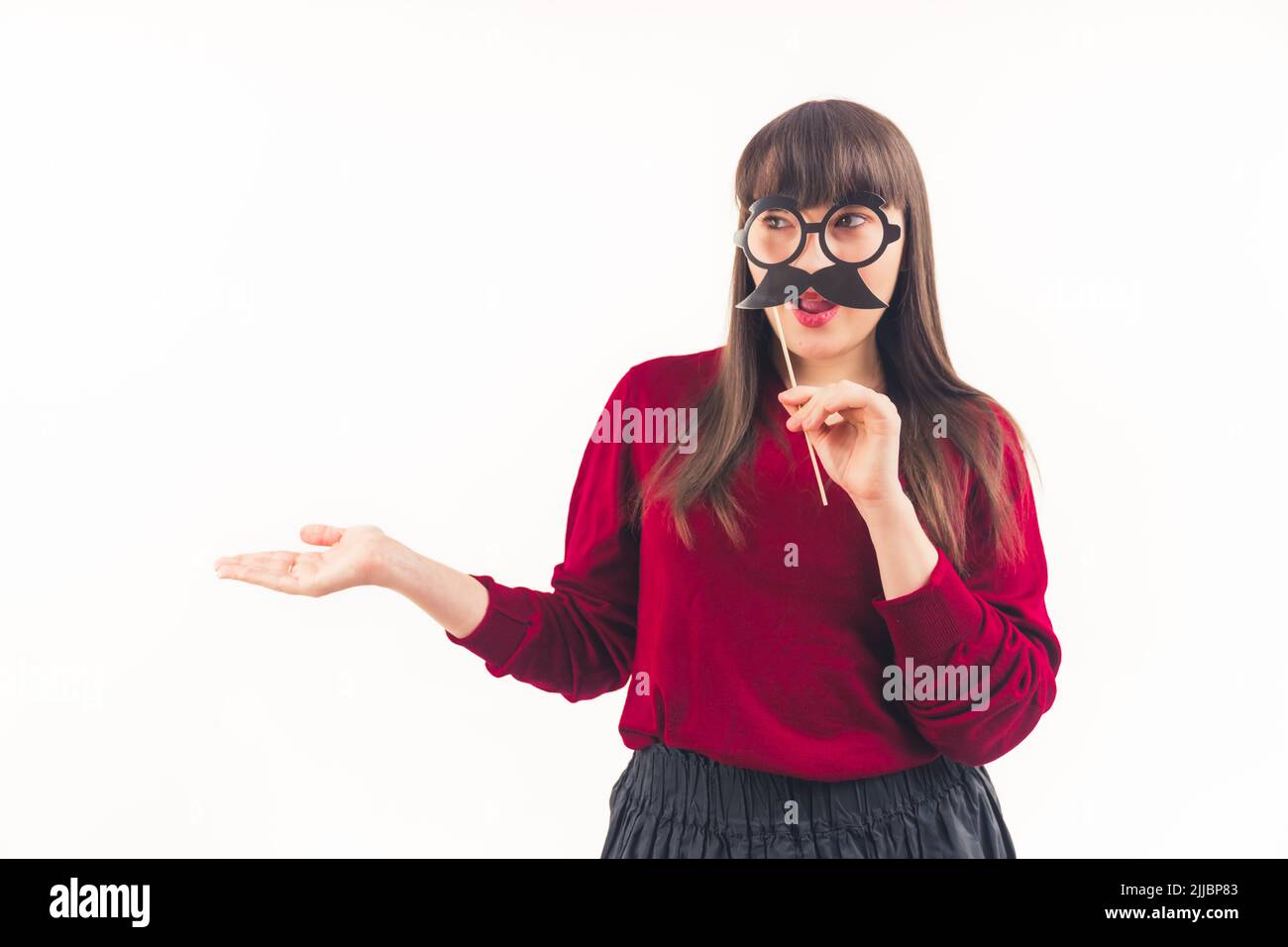 Femme brune caucasienne avec des bangs portant un chandail rouge tenant des lunettes de fausse moustache souriant. Prise de vue en studio. Espace de copie isolé. Photo de haute qualité Banque D'Images